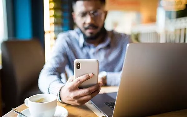 A man holding a smartphone writing a text message with a laptop in front of him